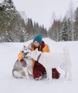A young woman is sitting in the snow next to her two husky dogs